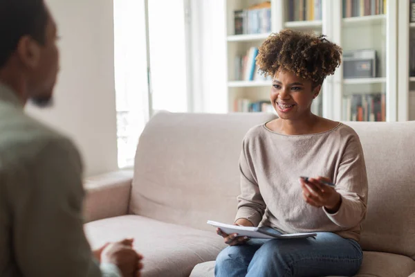Psychological help, support. Smiling friendly pretty young black woman in casual psychologist sitting on couch, holding folder, having conversation with patient african american man, copy space