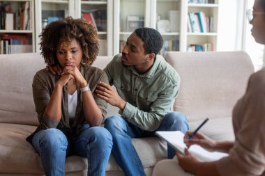 Attentive handsome young african american man husband hugging his upset depressed wife, black spouses visiting marriage counselor together. Problems in marriage, relationships concept