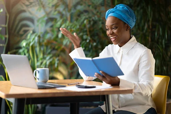Cheerful attractive millennial black woman business consultant have video call with client, sitting at table at cafe, smiling and gesturing at laptop webcam, reading data from notepad, copy space