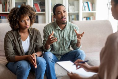 African american young husband and wife sitting on couch at family therapist office, fighting while visiting psychologist, emotional black man sharing his feeling with counselor, copy space