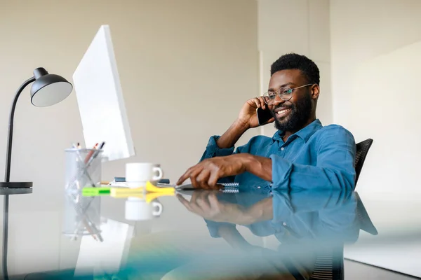 Positive black male entrepreneur talking on cellphone while working on modern computer in office. Businessman enjoying mobile communication, sitting at desk, copy space