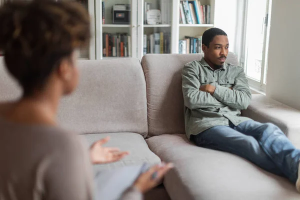 Reserved angry young black man resting on couch with arms crossed on chest, looking through the window at clinic, african american guy with addiction or PTSD attending psychotherapist woman