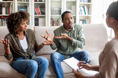 Emotional young black spouses fighting in front of family therapist, furious angry unhappy african man and woman sitting on couch, having conversation with psychologist. Marriage counseling concept