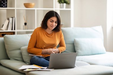 Sick Young Arab Woman Suffering Stomach Ache While Study With Laptop At Home, Upset Middle Eastern Female Having Menstrual Pain Or Problems With Digestion, Sitting On Couch And Touching Belly