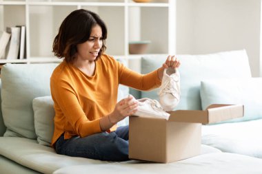Wrong Parcel. Anxious arab woman taking shoes out of box at home and frowning, confused middle eastern female suffering shipping mistake, feeling upset because of bad delivery service