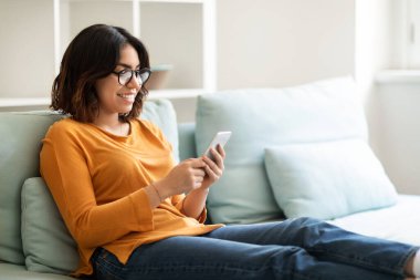 Smiling Middle Eastern Female In Eyeglasses Using Smartphone At Home, Portrait Of Happy Young Arab Woman Relaxing On Couch With Mobile Phone, Browsing Social Networks Or Shopping Online, Copy Space