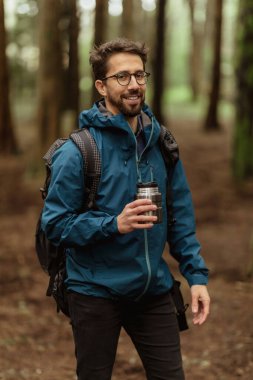 Glad millennial caucasian male tourist in glasses and jacket with backpack drink cup of hot coffee, resting in cold forest, enjoy adventure alone outdoor. Vacation, active lifestyle and camping, hiker