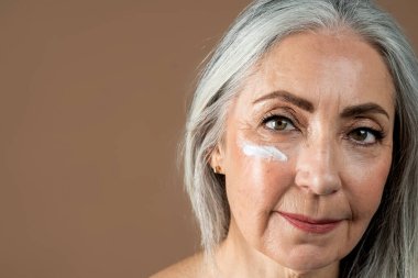 Calm old european lady with gray hair applies moisturizer on face, isolated on brown background, close up, studio. Beauty care, anti-aging and anti-wrinkle spa treatment, daily procedure with cream