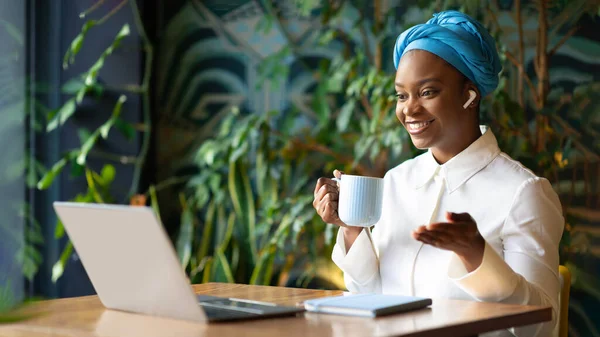 Friendly pretty young black woman in nice outfit independent contractor working from cafe, sitting at table, drinking coffee, using laptop and earpods, have video conference, panorama, copy space