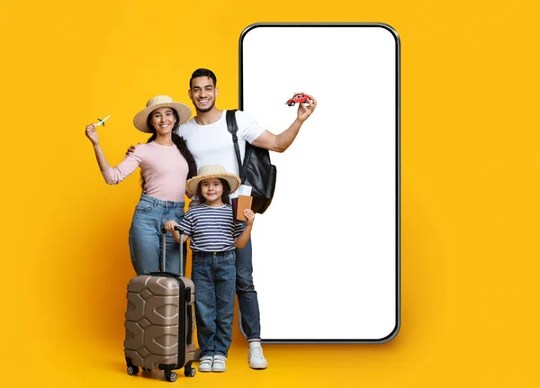 Family Travel Deal Concept. Happy middle eastern parents with little daughter holding plastic plane and car, posing by big phone with movkup on yellow background, mom, dad and child ready for vacation