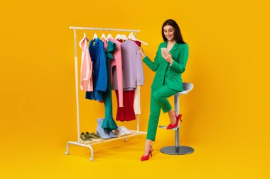 Mobile shopping. Happy lady using cellphone and purchasing clothes, sitting on chair near clothing rail over yellow studio background, full length