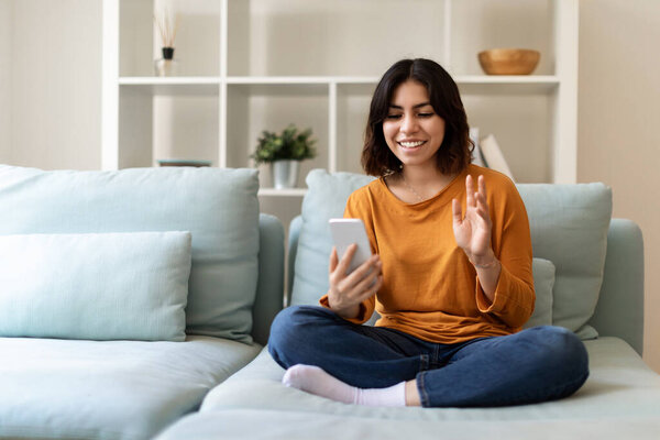Online Communication. Happy Arab Woman Making Video Call Via Smartphone At Home, Smiling Young Middle Eastern Female Sitting On Couch And Waving Hand To Mobile Phone Camera, Greeting Friend