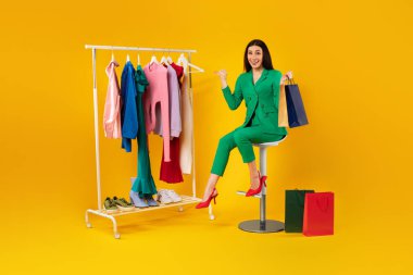 Shopaholic. Excited lady pointing at colorful clothes on clothing rail, shopping and choosing new outfil, posing on yellow background, full length shot