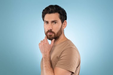 Attractive bearded middle aged man touching chin and looking at camera, posing over blue studio background. Male beauty and facial skincare routine
