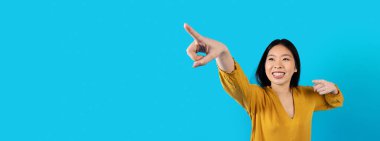 Excited happy pretty young asian woman in casual pointing at copy space for advertisement and smiling, isolated on blue studio background, panorama, web-banner. Nice deal, offer