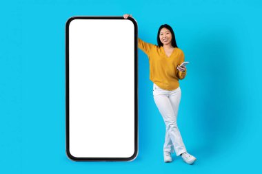 Happy cheerful smiling young chinese woman standing by big smartphone with white blank screen, showing nice online offer, using cell phone, blue studio background, full length, mockup