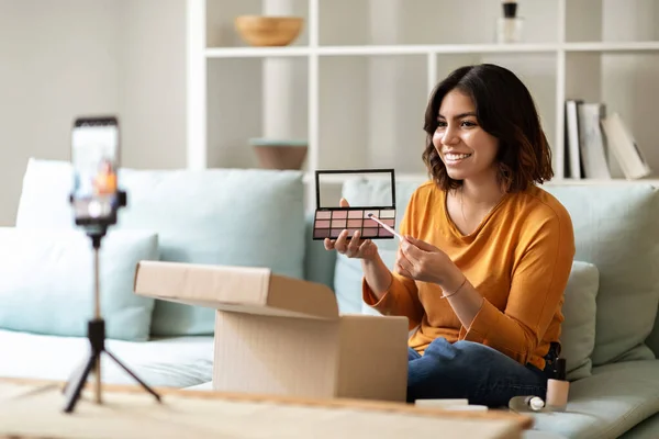 Unboxing Video. Beautiful Young Arab Woman Unpacking Parcel With New Cosmetics At Camera, Smiling Middle Eastern Female Demonstrating Eyeshadow Palette While Capturing Content For Her Beauty Blog