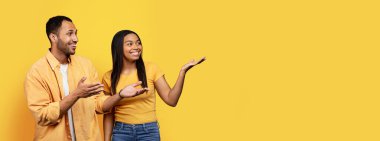 Smiling young african american man and female show hands on empty space, isolated on yellow background, studio, panorama. Relationship and love, recommendation and advice sale, news, ad and offer