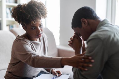 Friendly beautiful black woman therapist consoling upset young man, african american female psychologist touching sad crying patient shoulder, clinic interior. Psychological support concept
