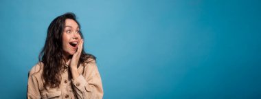 Cheerful excited shocked millennial european lady student with open mouth looking at empty space, isolated on blue background, studio, panorama. Reaction to surprise, good news emotions, ad and offer