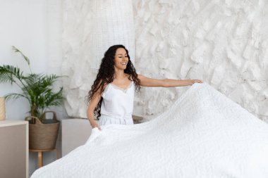 Cheerful millennial caucasian lady brunette in pajamas make bed, enjoy good morning, hold blanket in white bedroom interior, empty space. Domestic chores at weekend, free time, lifestyle and vitality