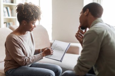 Attractive african american young woman psychologist having conversation with male patient, showing him charts, explaining mental processes. Black guy attending therapy session, copy space