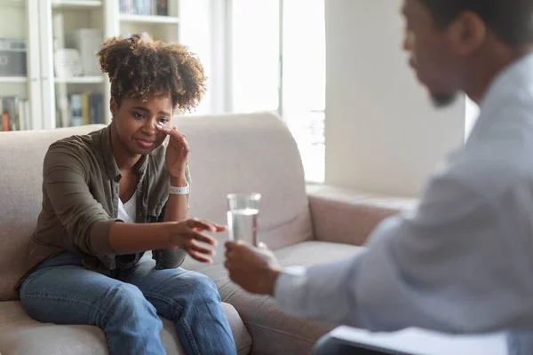 Black man in formal wear psychologist giving glass of water to crying devastated upset young african american woman. Millennial lady suffering from loneliness, grief, divorce visiting psychotherapist