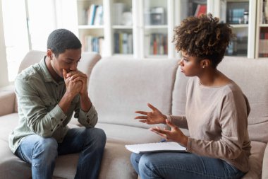 Frustrated pensive young african american man in casual outfit sitting on sofa at psychologist office, having therapy session. Black woman psychotherapist helping depressed guy with PTSD, copy space