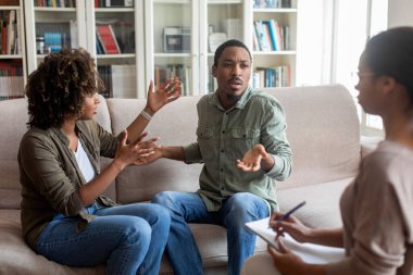 Hysterical angry emotional millennial black man and woman sitting on couch in front of female psychologist, gesturing and shouting, fighting during session at family therapist office, copy space