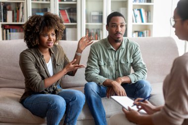 Angry attractive young black lady complaining about her husband at therapy session, unhappy married african american couple experiencing difficulties in relationships, visiting family counselor
