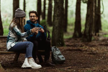Smiling millennial caucasian guy in jacket show smartphone to wife, sit on bench, relaxing in cold forest with backpack, enjoy adventure together outdoor. Map app for route vacation, active lifestyle