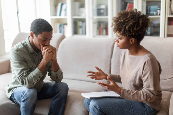 Frustrated pensive young african american man in casual outfit sitting on sofa at psychologist office, having therapy session. Black woman psychotherapist helping depressed guy with PTSD, copy space