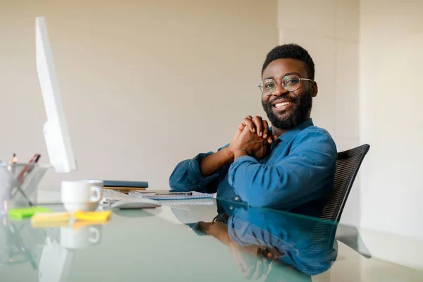 Millennial freelancer. Happy black man working with computer in office, enjoying remote work and distance job opportunities, sitting at desk and smiling at camera