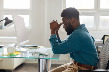 Thoughtful black male entrepreneur in glasses sitting at desk in front of computer, working online in office, side view. Pensive businessman thinking of problem solution