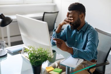 Tired black businessman feeling pain eyestrain, holding glasses and rubbing dry irritated eyes fatigued from computer work, man suffering from sight problem, sitting at workplace