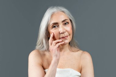 Calm old european lady with gray hair in towel touching face with fingers, looking at camera isolated on gray background, studio. Wrinkle treatment, skin care, cosmetology procedure, ad and offer