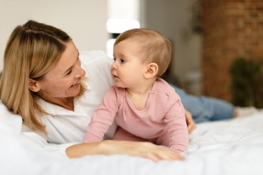 Loving mother playing with her cute baby daughter, kid crawling on bed and looking at mom, free space. Happy mommy enjoying motherhood routine and child care