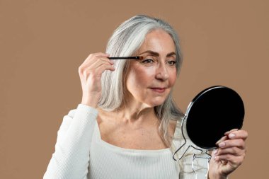 Smiling senior european female with gray hair applies mascara with brush on eyelashes, looks in mirror, isolated on brown background. Nude makeup, anti-aging cosmetics, beauty care, ad and offer