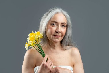 Calm aged european lady with gray hair hold bouquet of yellow flowers, enjoy spring, freshness, isolated on gray background, studio. Beauty procedures, skin care and spa treatments, ad and offer