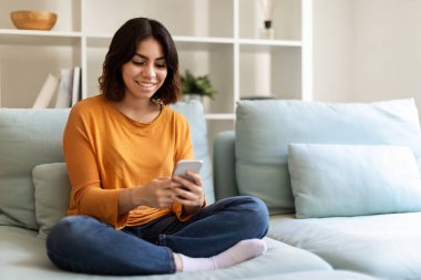 Smiling Young Arab Woman Resting With Smartphone On Couch At Home, Cheerful Middle Eastern Female Messaging With Friends Or Browsing Internet On Mobile Phone While Relaxing In Living Room, Copy Space