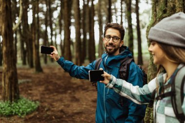 Glad millennial caucasian couple in jackets and backpack take selfie for blog on phone with empty screen in cold forest enjoy adventure together. Map app for route, vacation active, camping for hikers