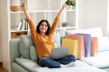 Black Friday. Overjoyed Arab Female With Laptop And Shopping Bags Celebrating Success While Sitting On Couch At Home, Happy Middle Eastern Female Raising Hands, Emotionally Reacting To Online Sales
