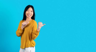 Studio shot of cheerful beautiful millennial asian lady in nice outfit posing on blue background, pointing at copy space and smiling, young chinese woman showing nice offer, panorama