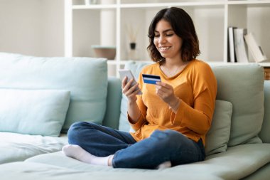 Mobile Payments. Portrait Of Young Arab Female Using Smartphone And Credit Card At Home, Smiling Middle Eastern Lady Sitting On Couch And Browsing App On Mobile Phone For E-Commerce, Copy Space