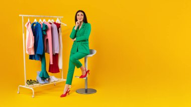 Style assistant. Young lady sitting near clothing rack with colorful clothes over yellow studio background, looking at copy space, panorama. Shopping and fashion business advertisement