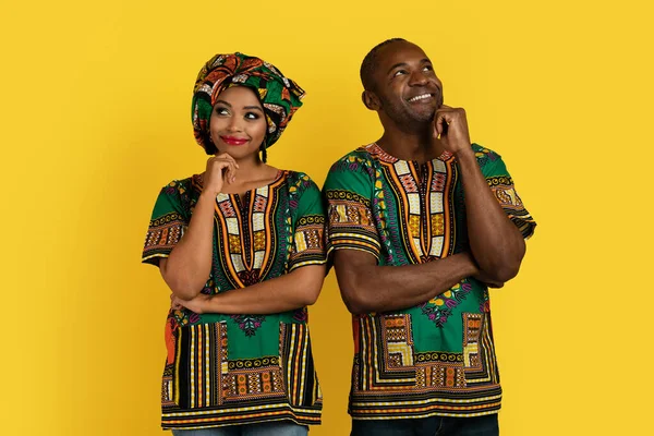 Excited attractive cheerful black man and woman in traditional african costumes posing over yellow studio background, touching chests and looking aside, thinking, making decision together