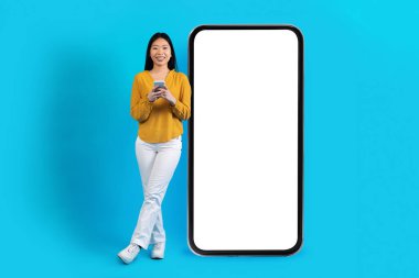 Positive cheerful pretty young korean lady in casual outfit posing next to big cell phone with white empty screen on blue studio background, using nice mobile app, mockup, copy space