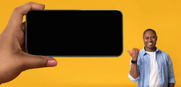 Cheerful Black Man Pointing At Big Blank Smartphone In Giant Female Hand, Smiling Young African American Male Showing Copy Space For Mobile Advertisement, Standing On Yellow Background, Mockup