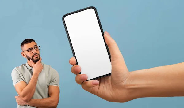 Pensive Arab Man Looking At Huge Blank Smartphone In Giant Female Hand, Big Arm Showing Empty Cellphone With White Screen To Thoughtful Middle Eastern Guy Standing Over Blue Background, Mockup