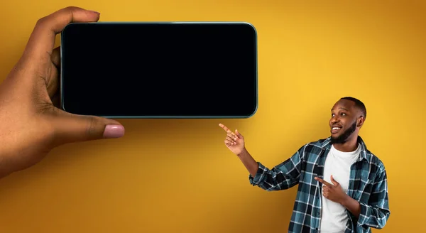 Positive Black Man Pointing Aside At Huge Female Hand With Blank Smartphone, Happy African American Guy Demonstrating Copy Space For Mobile App Or Website Design, Posing On Yellow Background, Mockup